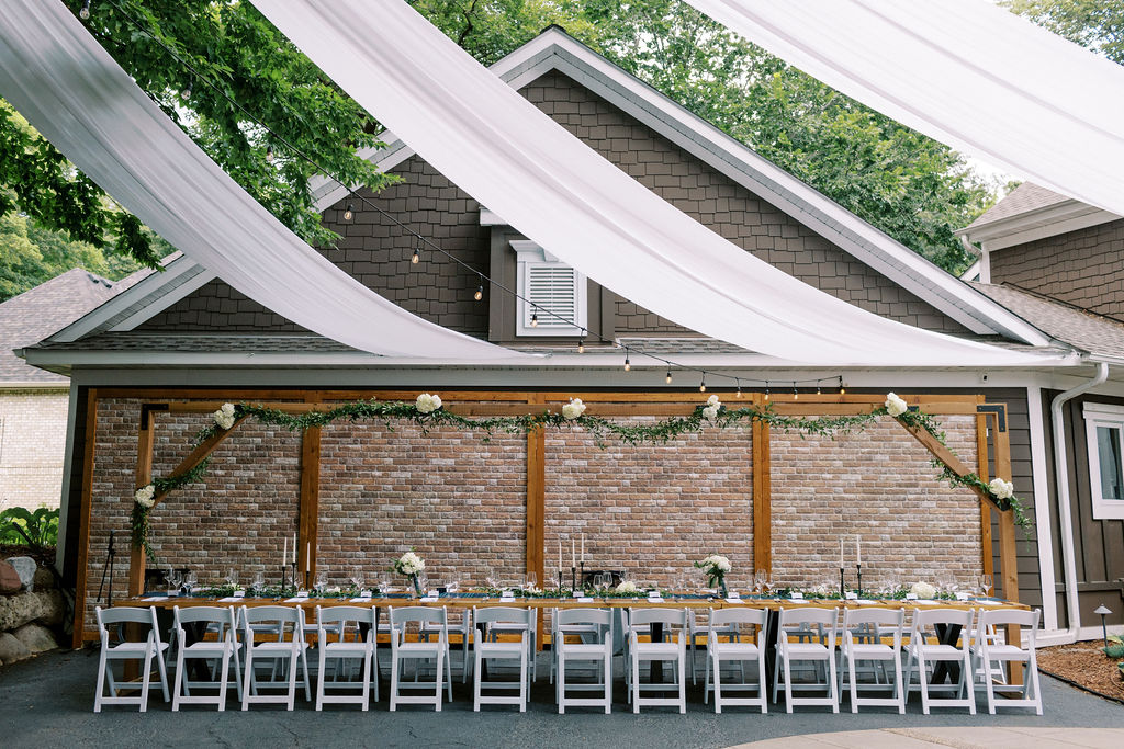 A long harvest table with tartan runners, white flowers and taper candles beneath draped white fabric and string lights in front of a brick wall